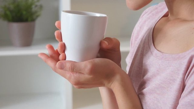 Young beautiful girl with short hair sit on bed in light room and enjoy drinking tea or coffee at morning at home.