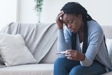 Depressed black girl holding pregnancy test, upset with positive result - obrazy, fototapety, plakaty