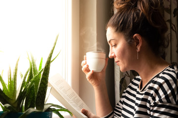 Adult Caucasian woman sitting near window at home relaxing in her living room reading book and drinking coffee or tea