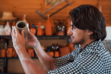 Young ceramist holds fresh handmade pot in hand and looks at results of his work