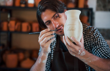 Young ceramist holds his fresh handmade pot and makes some detalizing by special tool stick