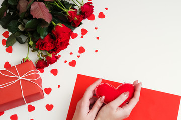 Valentine day. Top view of lady hands holding felt heart over blank greeting card. Red roses, gift box on white background.
