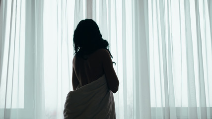 Romantic morning. Back view of nude brunette woman wrapped in white blanket, standing at window with sheer curtains. - obrazy, fototapety, plakaty
