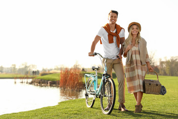 Obraz premium Young couple with bicycle and picnic basket near lake on sunny day