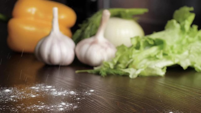 Flour is poured onto a wooden table on which are common items such as garlic, salad, pepper and onions. Slow motion view