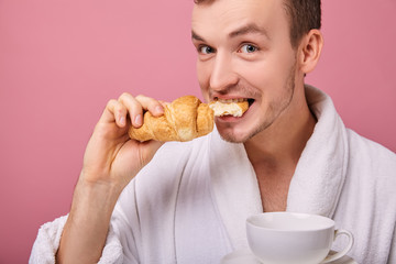 Funny pretty guy is biting fragrant croissant and holding white cup