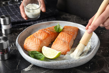 Woman adding sauce to cooked red fish on dark marble table, closeup
