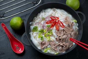 Pho bo soup with beef meat served in a cast-iron pan, elevated view over black stone background