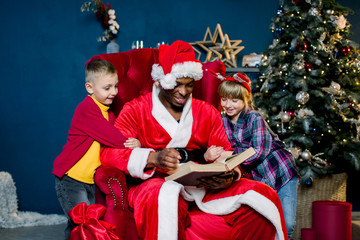 Small beautiful children, girl and boy, listen carefully to the story Santa Claus reads while sitting on a red chair on the background of a Christmas tree.