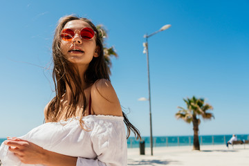 girl with hair fluttering in the wind looks questioningly through pink glasses, opening her mouth against the background of the beach and palm trees