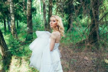 Portrait of young blond bride in forest with dress in hands