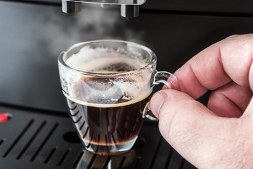 freshly brewed coffee is poured from the coffee machine into glass cup. Hand holding Cup