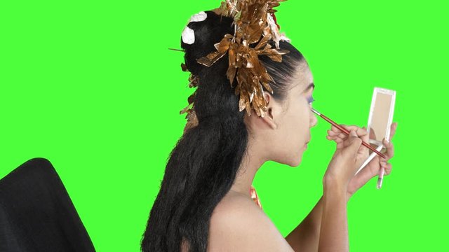 Balinese dancer coloring her eyelids while holding a small mirror in the studio. Shot in 4k resolution with green screen background