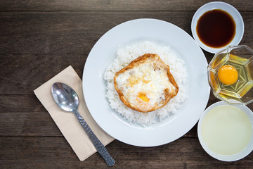 A fried egg is placed on the rice and put on a white ceramic dish. Vegetable oil and seasoning sauce and egg. On wooden table. close up shot. Top view.
