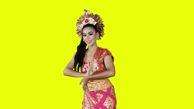 Attractive balinese dancer showing a welcoming gesture while wearing traditional costume in the studio. Shot in 4k resolution with yellow background