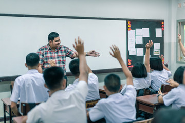 Fototapeta na wymiar An smiling Asian male high school teacher teaches the white uniform students in the classroom by asking questions and then the students raise their hands for answers.