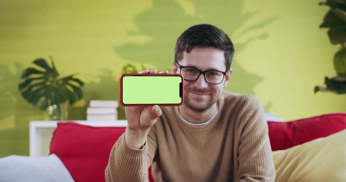 Young man holding mock-up smartphone greenscreen in horizontal position smiling showing modern internet app relaxing on sofa in home apartments.