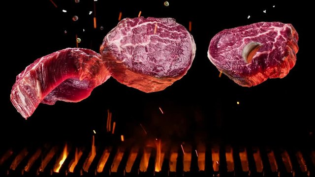 Raw pieces of steak with spices and salt slowly fall over a barbecue grill, coals burn below.