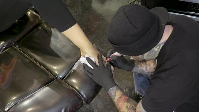 Tattoo artist placing small black and white tattoo on ladies foot Wide shot