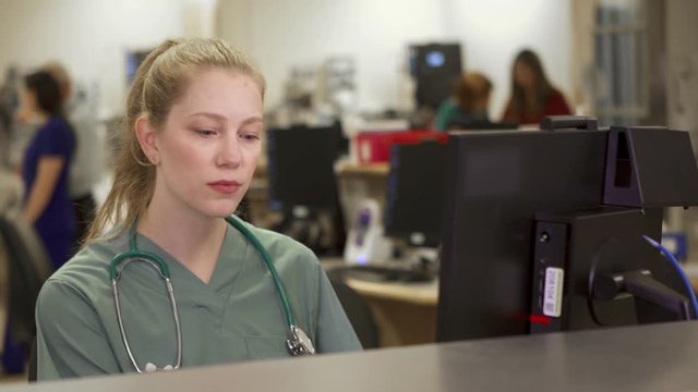 A Beautiful Young Blonde Nurse Receives A Phone Call And Smiles While Working At A Busy Nurses Station In The Hospital.