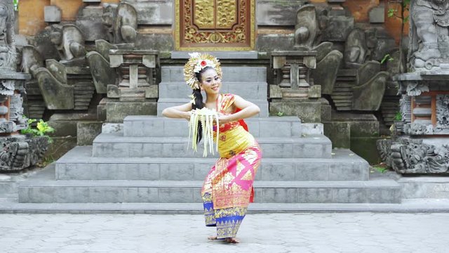 Slow motion of balinese dancer showing a welcoming gesture while holding frangipani flower and wearing traditional costume in the temple