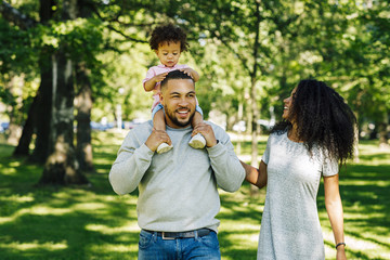Obraz premium Two parents and their son walking outside. Mother looking on her son sitting father's shoulders.