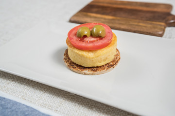Minimal spanish tapa on a white plate. Potato omelette with tomato, olive on a bread base. Close up image.