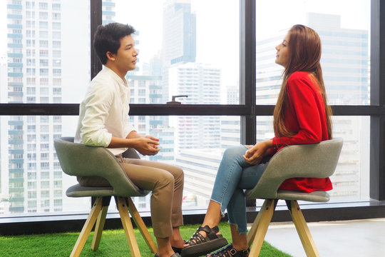 Side View Of Two Young People Sitting On The Chairs In The Office Facing Each Other Talking And Discussing Information On Work And Life Problems. Casual Meeting And Interview And Lifestyle Concept
