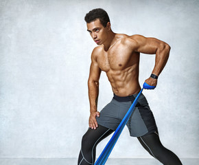 Strong man working with resistance band. Photo of man with athletic body on grey background....