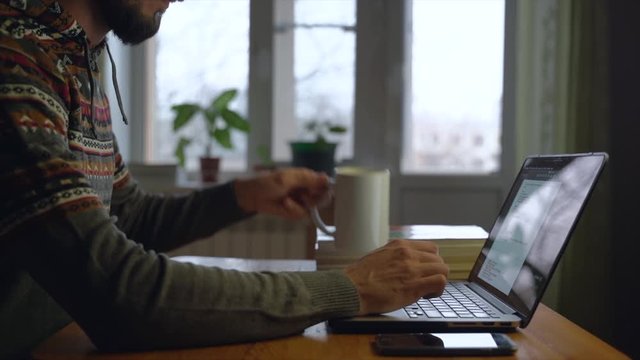 Bearded freelancer is drinking a tea in front of laptop at his home while working.