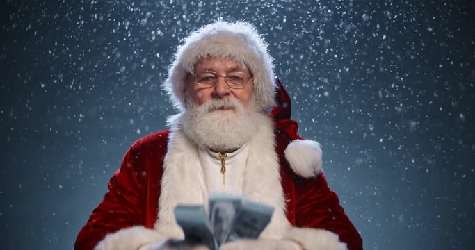 Santa clause looking at camera and showing packs of money in his hands, isolated on blue snowy background - christmas spirit concept close up 4k footage