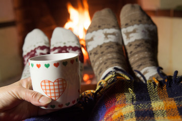 Man and woman in warm knitted socks with cups of hot punch in front of the fireplace