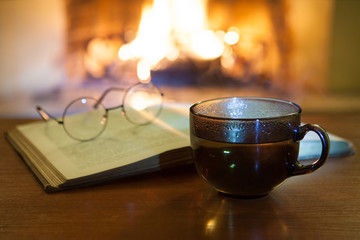 An open ancient book, vintage glasses, a cup and biscuits in front of the fireplace. Evening fairy tales. Fantasy.