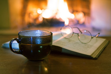 An open ancient book, vintage glasses, a cup and biscuits in front of the fireplace. Evening fairy tales. Fantasy.