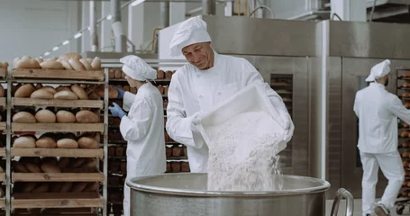 Poster Grote bakker in de bakkerij-industrie bereidt het deeg voor, voegt de bloem toe in een grote container. Achtergrondarbeiders schikken het brood en transporteerden het gebakken brood. © VisionPro
