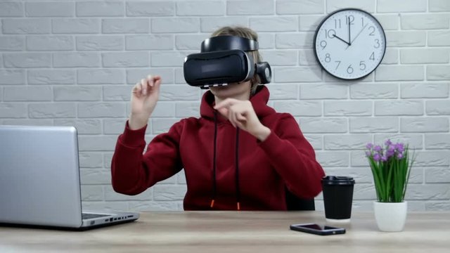 Girl in headset of virtual reality on head touch something like push on button or pointing at floating virtual screen work at desk with laptop