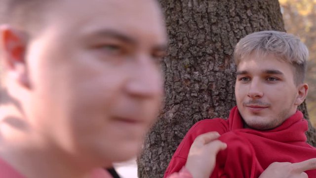 Young caucasian man in pink pullover rudely grabbs another guy in red hoody with dyed hair, by the shirtfront, . Then second one points to somewhere with a finger. Autumn park or forest outdoor.