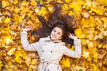 Young beautiful little girl in beige coat lying on yellow leaves
