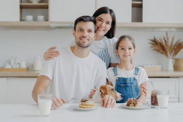 Obraz na płótnie Canvas Happy family and dog pose in cozy kitchen, eat fresh homemade pancakes with chocolate and milk, look positively at camera. Mother in apron embraces husband and daughter, likes cooking for them