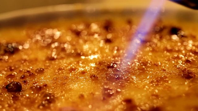 Caramelized sweet dessert fire cooking. French cuisine - creme brulee cooking process using burner flame. Selective focus. Macro shot. 