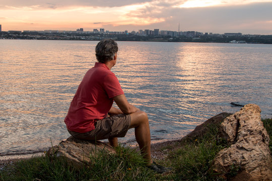 Mature Man Hanging Out At The Park Wearing Casual Clothes Siting On A Rock Watching The Sunset