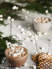 Two cup with hot cocoa and marshmallows on a white wooden table. Nearby are fresh waffles and marshmallows. The background is decorated with coniferous branches. New Year's breakfast concept, close-up