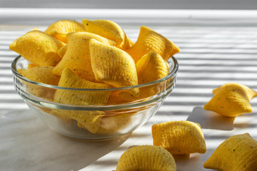 Corn chips with the taste of sour cream and onions in the form of pillows lie in a glass bowl on a white background, consecrated by the sun's rays passing through the closed blinds