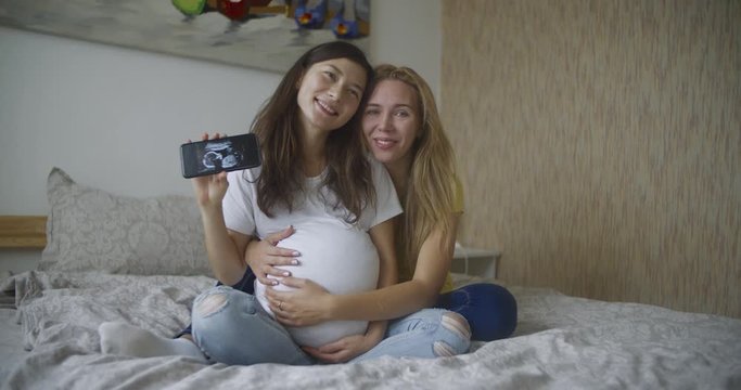 Pregnant multinational lesbian couple shows at an ultrasound image of his baby on the phone. LGBT