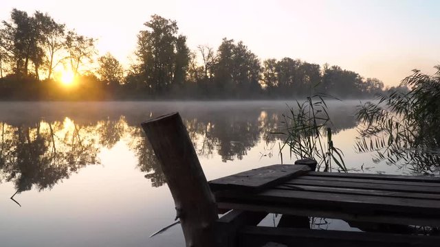 Sunrise on the riverbank. Landscape with wooden fisher bridge, reeds, rising sun and smoke on the water.