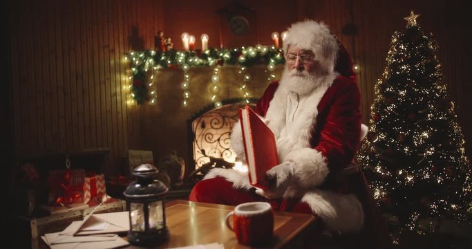 Joyful santa clause sitting in his rocker in decorated room, reading a book with red cover - holiday mood, christmas spirit concept 4k footage