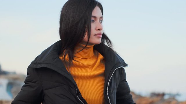 Medium shot of attractive pensive Asian girl in down jacket thoughtfully walking alone on seaside