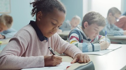 In Elementary School Classroom Brilliant Black Girl Writes in Exercise Notebook, Taking Test and Writing Exam. Junior Classroom with Diverse Group of Children Working Diligently and Learning New Stuff