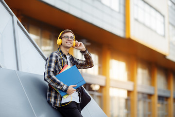 Handsome young male student in casual outfit with yellow headphones holding folders while listening...