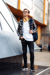 Full lengh photo of young male student in casual outfit with yellow headphones and blue backpack standing on the street.